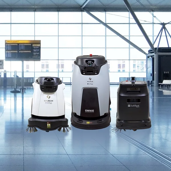 3 autonomous floor cleaning robots of different sizes standing on a polished airport terminal floor, with a flight information board behind them