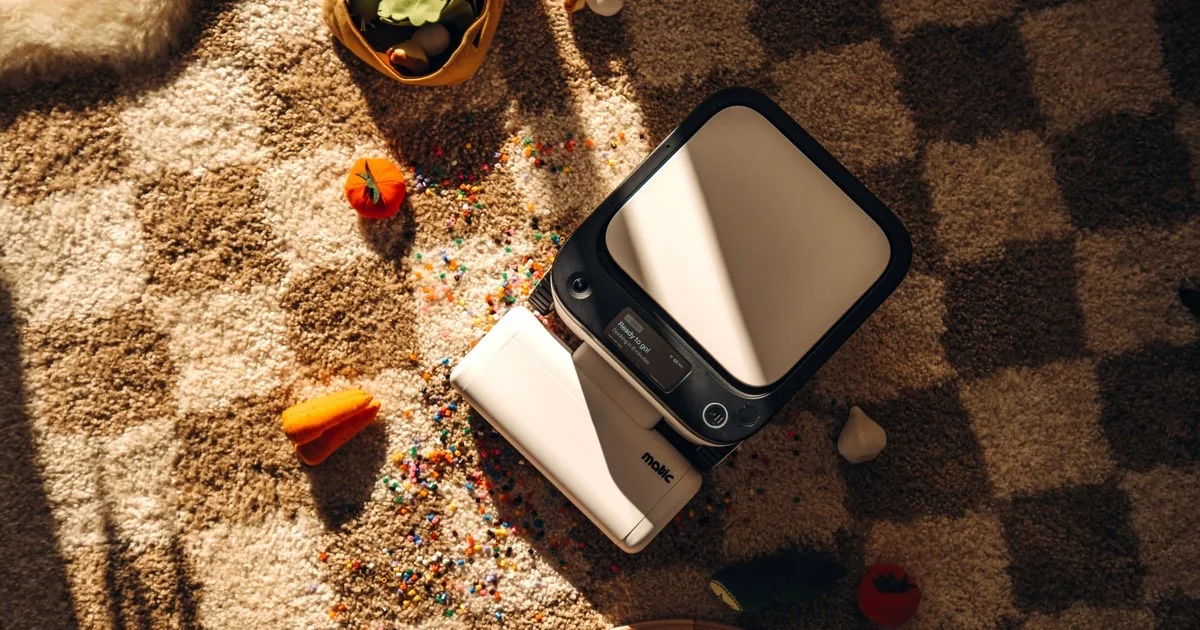 A Matic floor cleaning robot seen from above on a checkered rug, vacuuming up spilled craft beads next to toy vegetables