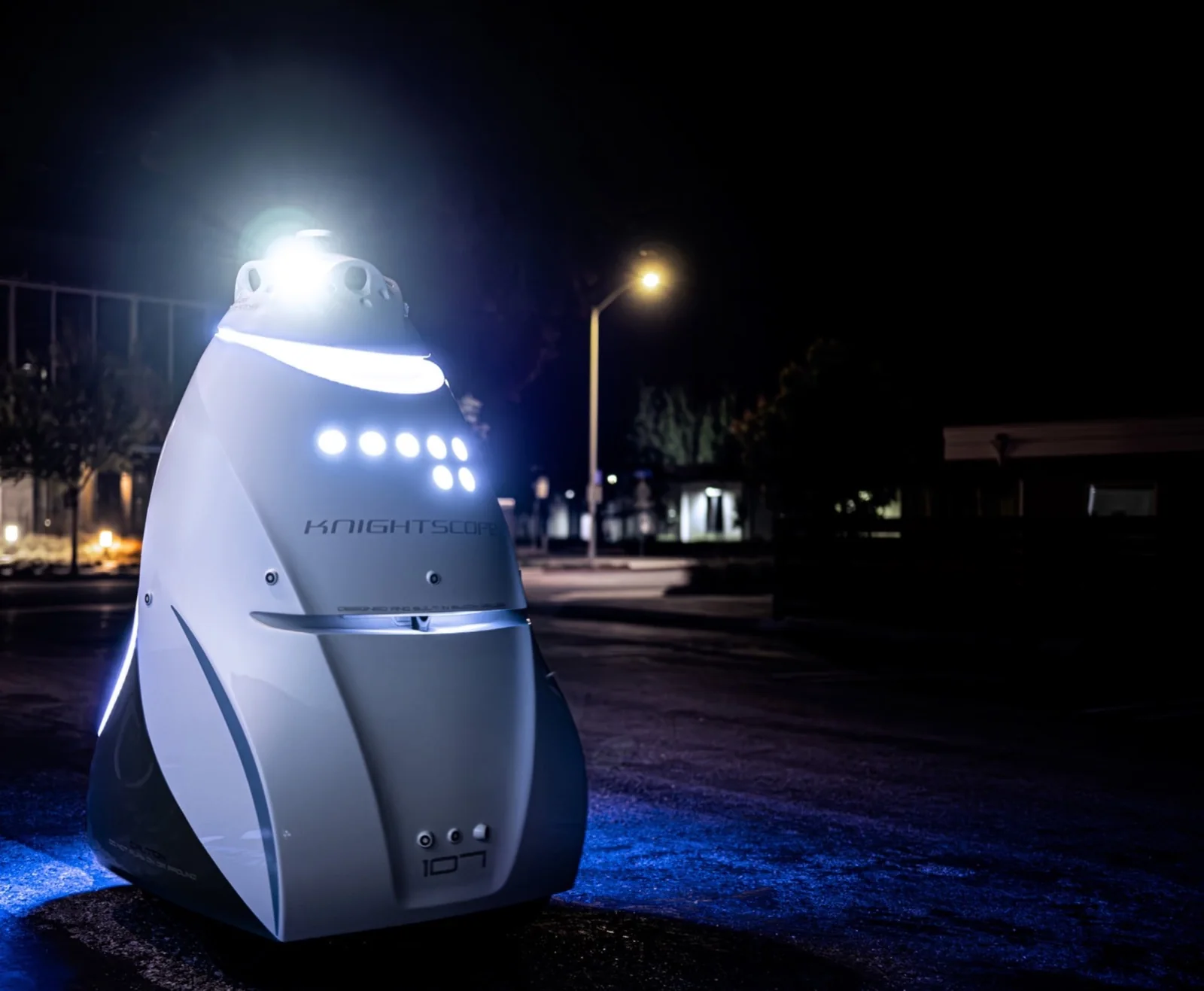 A white Knightscope K5 security robot lit up at night in a parking lot, with a street lamp behind it