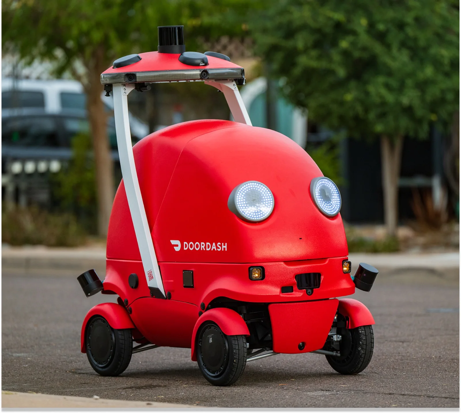A red DoorDash Dot delivery robot on 4 wheels, with round LED headlights and a sensor mast on top, parked on a paved street with trees behind it